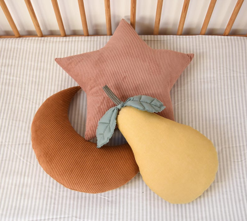 May include: Three plush pillows in the shape of a star, a crescent moon, and a pear. The star is pink corduroy, the moon is brown corduroy, and the pear is yellow.