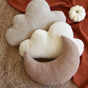 May include: Three plush pillows in the shape of clouds and a crescent moon. The pillows are in shades of brown and white. The pillows are on a woven rug.