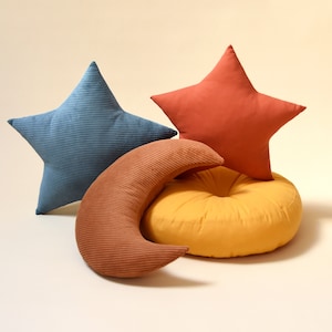 Op de afbeelding: Drie decoratieve kussens in de vorm van een ster, een halve maan en een rond kussen. Het stervormige kussen is rood, het halve maankussen is bruin en het ronde kussen is geel.
