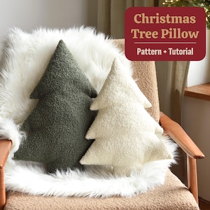 May include: Two Christmas tree-shaped pillows, one olive green and one cream, rest on a chair covered with a white faux fur throw. The pillows have a textured, plush appearance. A sign in the background reads "Christmas Tree Pillow Pattern + Tutorial."