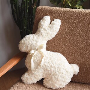May include: A white plush bunny rabbit with a white satin bow. The bunny is sitting on a brown couch.