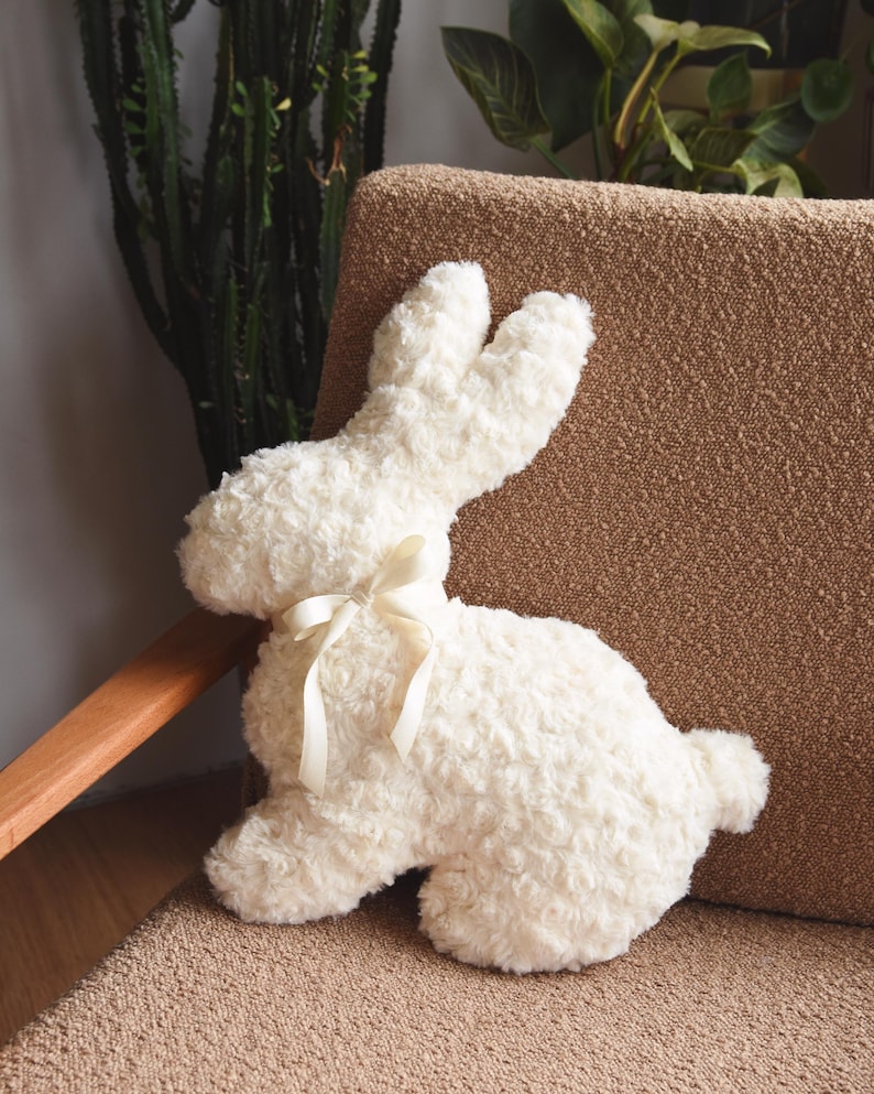 May include: A white plush bunny rabbit with a white satin bow. The bunny is sitting on a brown couch.