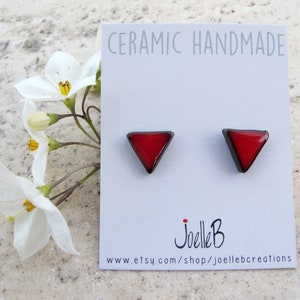 Puede incluir: Un par de pendientes hechos a mano de cerámica. Cada pendiente es un triángulo rojo con un borde oscuro. Los pendientes se muestran en una tarjeta blanca con el texto "CERAMIC HANDMADE" y el nombre del artista, "Joelle B".