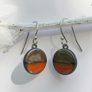 Peut inclure: Une paire de boucles d'oreilles pendantes en argent avec des breloques rondes en céramique. Les breloques sont bicolores, avec un dessus gris foncé et un dessous orange vif.