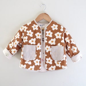 May include: A brown quilted jacket with white daisy print and gingham pockets. The jacket has a button closure and is lined with a white fabric.