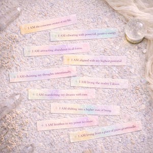 Puede incluir: Tarjetas de afirmación de colores pastel con frases como "YO SOY el creador consciente de mi vida" y "YO SOY atrayendo la abundancia en todas sus formas". Las tarjetas están dispuestas sobre una superficie clara y texturizada, con una bola de cristal y cristales en el fondo.