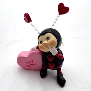 May include: A whimsical figurine of a ladybug-like character with a black body and red heart accents. It holds a pink heart-shaped candy with "BEE MINE" text. The figurine has spring antennae with red heart toppers and is posed in a thoughtful manner.