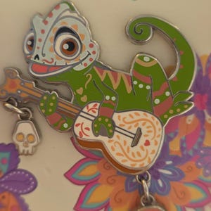 May include: A colorful enamel pin featuring a green chameleon playing a guitar, with a skull charm. The pin is set against a floral background and includes the text "Pin Trading Day 2018" and "Edition Limitée 500 Exemplaires".
