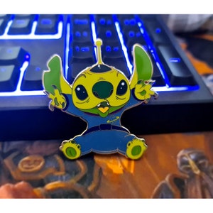 Puede incluir: Un pin de esmalte verde y azul con un personaje de dibujos animados extraterrestre con ojos grandes y una sonrisa de dientes. El personaje lleva un traje espacial azul con un diseño de planeta en el pecho.