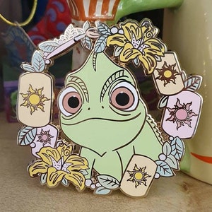以下が含まれることがあります： 大きな目をした緑色のカメレオンが、黄色い花とピンクと黄色の太陽模様の提灯の輪で囲まれている、金色のエナメルピンです。
