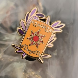 Può includere: Uno spillo dorato con un designo viola e oro. Lo spillo presenta un sole stilizzato con un centro rosso e le parole "Wish" e "Upon" in un font corsivo.