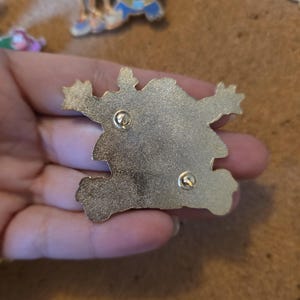 Pu&ograve; includere: Una spilla in smalto color oro con un design a mostro dei cartoni animati. La spilla ha due fermagli per un fissaggio sicuro. Altre spille sono visibili sullo sfondo.