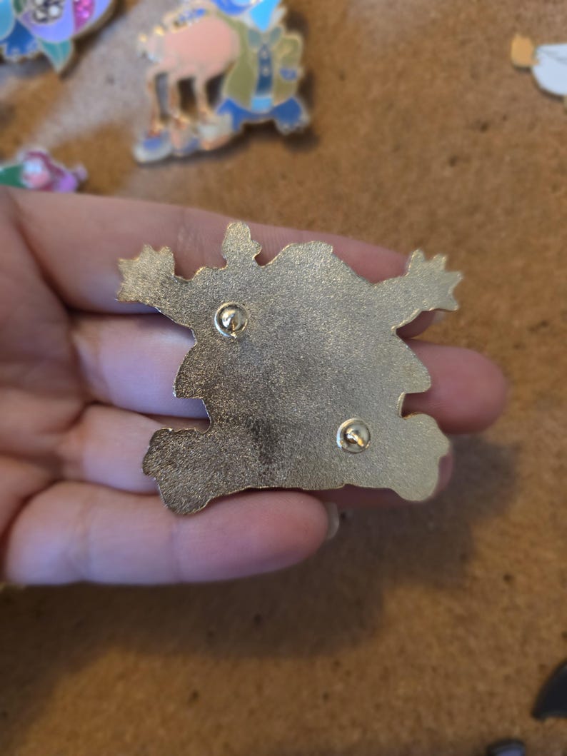 Pu&ograve; includere: Una spilla in smalto color oro con un design a mostro dei cartoni animati. La spilla ha due fermagli per un fissaggio sicuro. Altre spille sono visibili sullo sfondo.