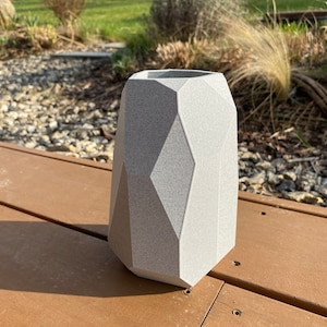 May include: A gray geometric ceramic vase with a textured surface. The vase is sitting on a wooden surface.