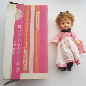 May include: A vintage doll with brown curly hair, wearing a pink dress with white polka dots and a white apron. The doll is holding a small white purse and is wearing black shoes. The doll is standing on a white surface. The doll is in its original box, which is pink and white and has the text "Horsman" on it.