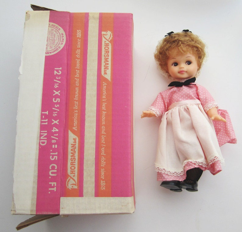 May include: A vintage doll with brown curly hair, wearing a pink dress with white polka dots and a white apron. The doll is holding a small white purse and is wearing black shoes. The doll is standing on a white surface. The doll is in its original box, which is pink and white and has the text "Horsman" on it.