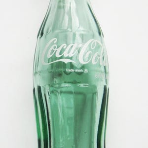 May include: A vintage green Coca-Cola glass bottle. The bottle has the Coca-Cola logo in white script and the words "Trade-mark ®". The bottle is empty and has a classic, curved shape.
