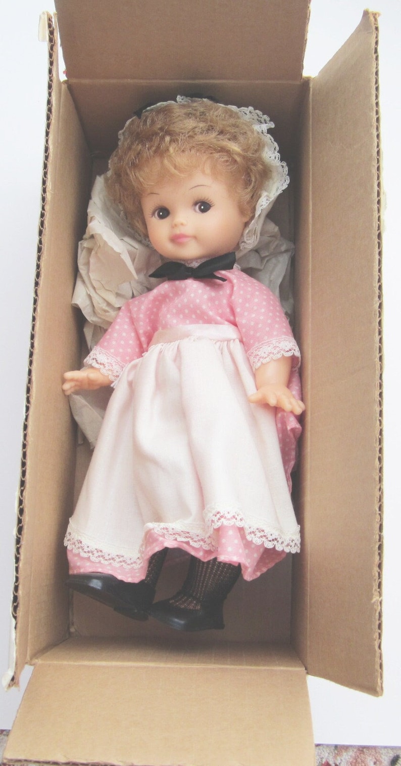 May include: A pink and white doll with brown hair and blue eyes is wearing a pink dress with white polka dots and a white lace trim. The doll is in a cardboard box.