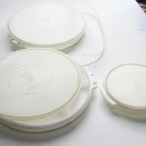 May include: A collection of off-white, round plastic food storage containers with lids. The lids have a slightly translucent appearance and are stacked together. The containers are of varying sizes, with handles on the sides.
