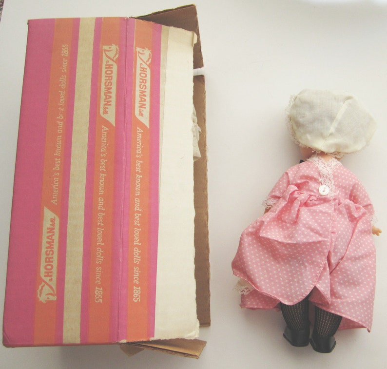 May include: A pink and white polka dot dress with a white lace trim and a white bonnet. The dress is on a box with the text "HORSMAN America's best known and best loved dolls since 1865" printed on it.