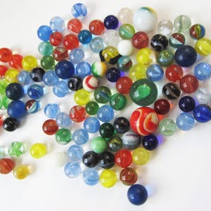 May include: A large collection of colourful glass marbles in various shades of blue, green, red, yellow, and white. The marbles are arranged in a loose pile on a white surface.