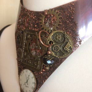 Op de afbeelding: Een koperkleurige, driehoekige chokerketting met een steampunk-ontwerp. De ketting is versierd met verschillende tandwielen, wijzerplaten en andere metalen versieringen. De wijzerplaat geeft "Paris" aan.