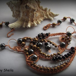 May include: A copper wire necklace with black and gold beads. The necklace is wrapped in a spiral pattern and has a delicate, intricate design. The necklace is displayed on a white surface with a seashell.