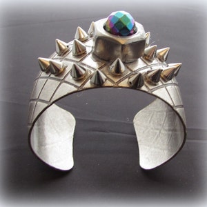 Może przedstawiać: Srebrna bransoletka z siatką i kolcami. Bransoletka ma dużą srebrną nakrętkę z wielokolorowym, tęczowym kamieniem na górze.