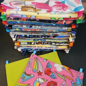 May include: A stack of colourful fleece fabric squares with different patterns, including a sloth, hearts, and rainbows. The fabric squares are tied together with colourful ribbons. A pink and green fleece fabric square with a rainbow, hearts, and stars pattern is in the foreground.