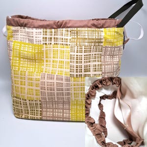 Puede incluir: Una cesta de almacenamiento rectangular de tela con un diseño de patchwork en amarillo, verde oliva y topo. La cesta tiene un interior y asas marrones, con un cierre de cordón blanco. El diseño presenta un patrón de cuadrícula.