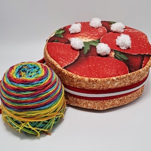 Puede incluir: Una caja redonda cubierta de tela con un diseño de fresas y pompones blancos. Una madeja de hilo colorido está al lado de la caja. La caja tiene una cinta roja y blanca. El hilo es una mezcla de amarillo, verde, azul y rojo.
