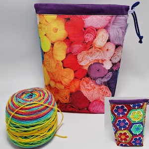Op de afbeelding: Een kleurrijke garen opbergtas met een paarse rand en trekkoord. De tas heeft een levendige print van verschillende garenkleuren. Een bol regenboogkleurig garen ligt naast de tas en toont een verscheidenheid aan tinten.