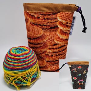 Puede incluir: Una bolsa de proyecto alta de tela marrón con estampado de gofres, cierre de cordón y una pequeña bola de hilo colorida. También se ve una bolsa más pequeña con la parte superior marrón y un estampado de coches, conejitos y flores.