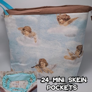 Puede incluir: Una bolsa de tela azul claro con una parte superior marrón y una cremallera. La bolsa tiene un estampado de ángeles en nubes. La bolsa tiene 24 mini bolsillos y el texto "24 MINI SKEIN POCKETS" está impreso en la parte delantera.