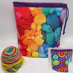 Puede incluir: Una bolsa grande para guardar hilo con un estampado vibrante de bolas de hilo en arcoíris. La bolsa tiene una parte superior morada y cierre de cordón. También se ve una bolsa más pequeña con un diseño de ganchillo y una bola de hilo arcoíris.