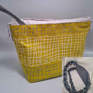 Puede incluir: Un bolso tote amarillo con un patrón de cuadrícula blanca y un asa gris. El bolso tiene un cierre de cordón y un interior de color claro. También se muestra una diadema gris a juego.