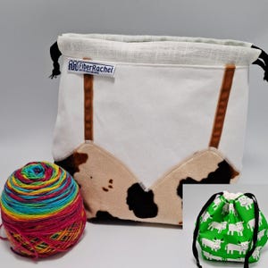 Può includere: Una borsa porta-progetti bianca con chiusura a coulisse, inserti in finta pelliccia con stampa mucca marrone, cordini neri ed etichetta FiberRachel. Accanto si trova un gomitolo di lana arcobaleno multicolore. Un riquadro mostra una borsa verde con mucche.