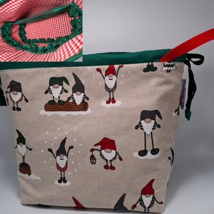 Puede incluir: Una bolsa de cordón verde y roja con una tela beige que presenta un patrón de gnomos. La bolsa tiene un cordón negro y un asa roja. La bolsa está forrada con tela verde y tiene un patrón de cuadros rojos y blancos.