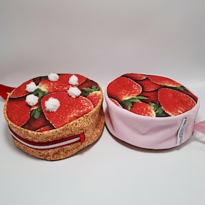 Puede incluir: Dos bolsas redondas de tela. Una está diseñada para parecerse a un pastel de fresas, con un lado marrón y beige, una parte superior con estampado de fresas rojas, pompones blancos y una cremallera roja. La otra es rosa.