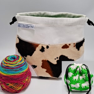 Può includere: Una borsa con coulisse con una sezione superiore bianca e una sezione inferiore con stampa mucca marrone, nera e bianca, con un'etichetta FiberRachel. Un gomitolo di lana multicolore è accanto e un riquadro mostra una borsa verde con mucche.