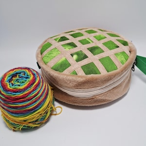 Puede incluir: Una bolsa redonda de hilo, de color beige, con un diseño de celosía verde y beige en la parte superior, junto a una bola de hilo colorida. La bolsa tiene cierre de cremallera y un asa verde. El hilo es una mezcla de colores arcoíris.