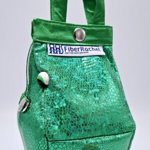 Puede incluir: Una bolsa de tejido de punto verde con una  etiqueta "FiberRachel Knitting Bags and More". La bolsa tiene un  diseño festoneado y un  acabado brillante y texturizado.