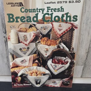 Op de afbeelding: Een magazinecover getiteld "Country Fresh Bread Cloths" van Leisure Arts. De afbeelding toont verschillende manden gevuld met brood, eieren en appels, elk gevoerd met geborduurde doeken. De doeken hebben verschillende ontwerpen en het woord "Bread" is zichtbaar.