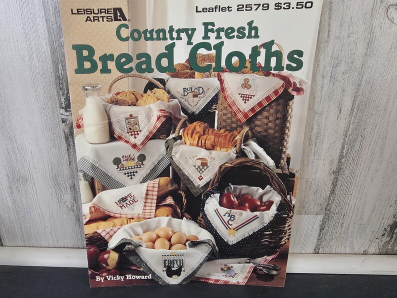 Op de afbeelding: Een magazinecover getiteld "Country Fresh Bread Cloths" van Leisure Arts. De afbeelding toont verschillende manden gevuld met brood, eieren en appels, elk gevoerd met geborduurde doeken. De doeken hebben verschillende ontwerpen en het woord "Bread" is zichtbaar.