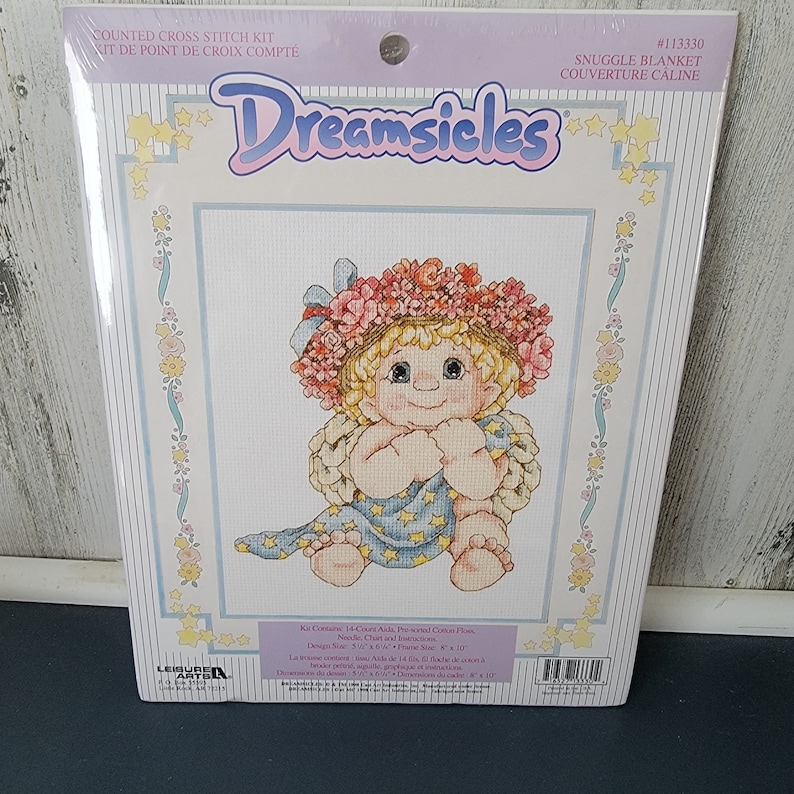 May include: A counted cross stitch kit featuring a cartoon baby girl wearing a flower crown and holding a blue blanket with stars. The kit includes 14 count Aida cloth, a needle, chart and instructions. The design size is 7.6 cm x 15.2 cm and the frame size is 20.3 cm x 27.9 cm.