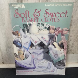 May include: A vintage craft book titled "Soft & Sweet Basket Cloths" by Leisure Arts. The cover features a pastel illustration of embroidered cloths, baskets, and a small table setting. The color palette includes soft pinks, blues, and whites.