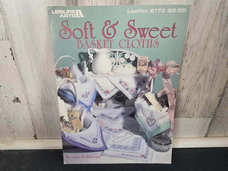 May include: A vintage craft book titled "Soft & Sweet Basket Cloths" by Leisure Arts. The cover features a pastel illustration of embroidered cloths, baskets, and a small table setting. The color palette includes soft pinks, blues, and whites.