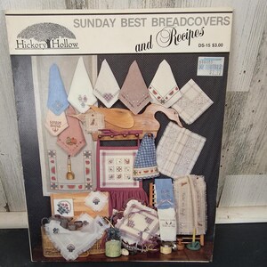 May include: A book titled "Sunday Best Breadcovers and Recipes" by Hickory Hollow. The cover displays various embroidered bread covers in pastel colors, including blue, pink, and white, with decorative patterns and designs. A price tag is visible.
