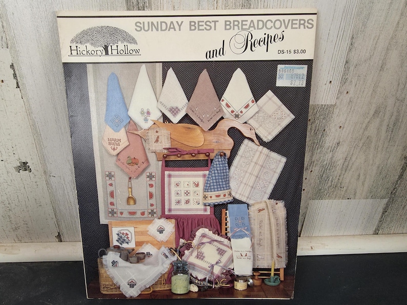May include: A book titled "Sunday Best Breadcovers and Recipes" by Hickory Hollow. The cover displays various embroidered bread covers in pastel colors, including blue, pink, and white, with decorative patterns and designs. A price tag is visible.