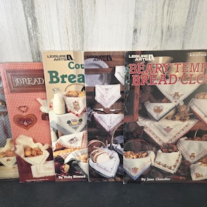 May include: A collection of vintage Leisure Arts booklets featuring bread-themed cross-stitch patterns. The covers display images of bread baskets, embroidered cloths, and the text "Bread" and "Beary Tempting Bread Cloths."
